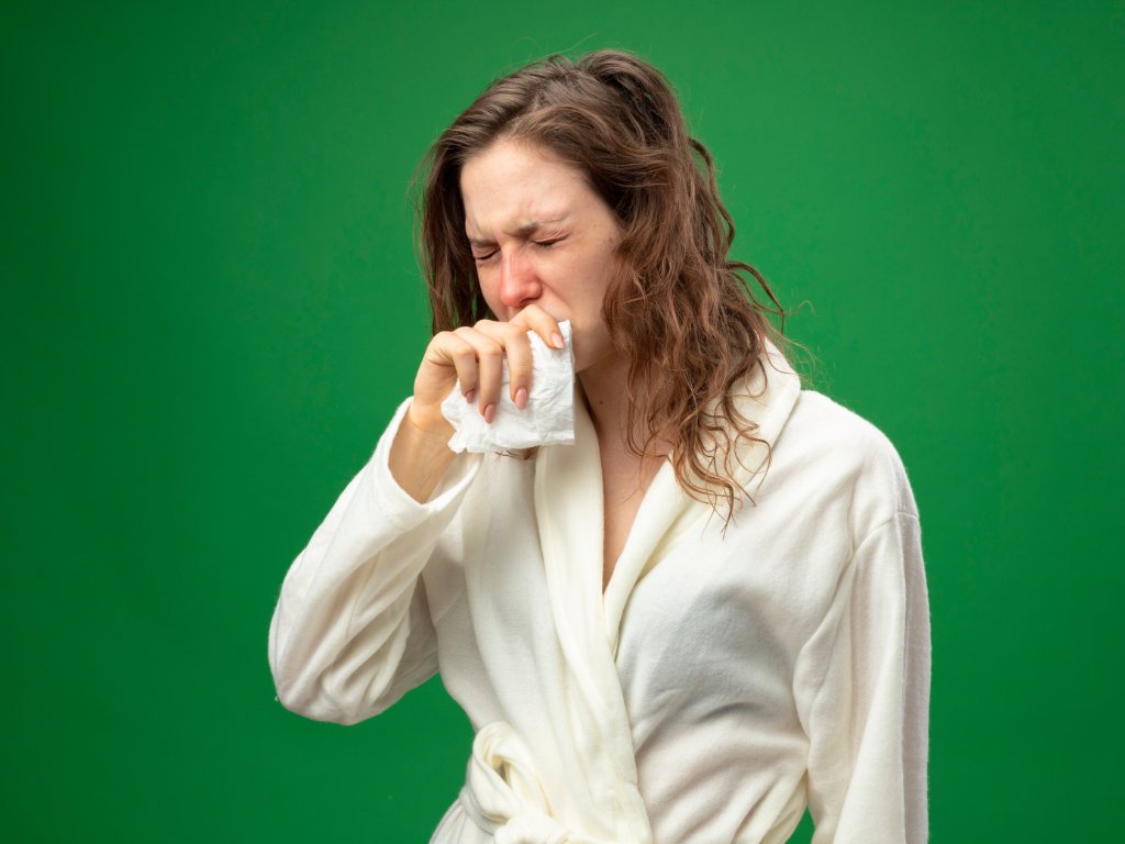 A sick woman in a white robe sneezing into a tissue, illustrating the need for immunity boosts during winter cold and flu season