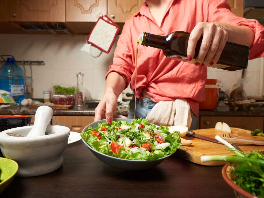 Person preparing fresh low-carb Mediterranean dishes by drizzling olive oil over a colorful salad with tomatoes and greens in a modern kitchen