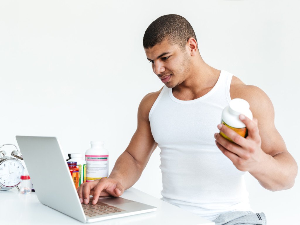 A man examining a pre-workout supplement bottle while researching on a laptop to avoid stimulant overload in pre-workout supplements