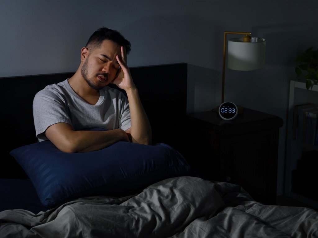 Man dealing with night wakings and poor sleep quality, sitting frustrated in bed at 2:33 AM with hand on forehead