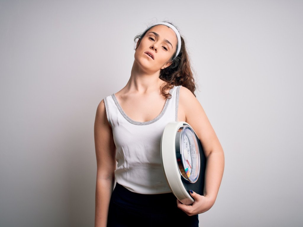 Woman looking frustrated while holding scale, representing the challenge to shed pounds when hitting weight loss plateaus