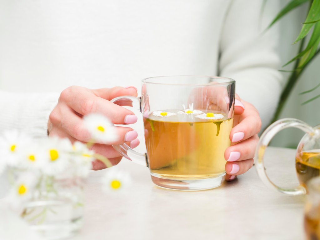 Hands holding warm chamomile tea with natural sleep aids properties, surrounded by fresh daisy flowers on white table
