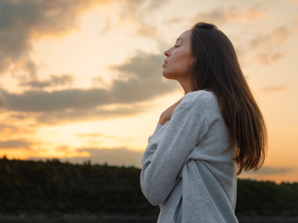 Woman experiencing mindful meditation benefits through consistent practice, standing peacefully with eyes closed during golden hour in natural setting