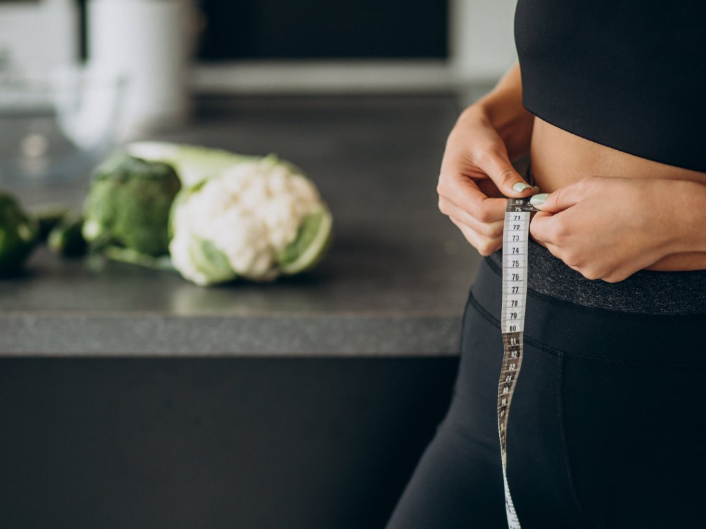 Person measuring waist with tape measure in kitchen with fresh vegetables showing how to track weight loss in your 40s and work with metabolic changes