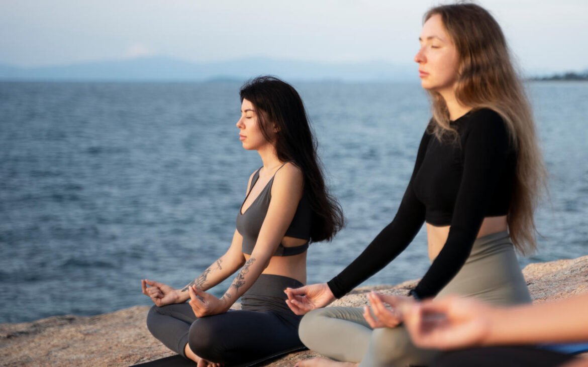 Two women practicing essential mindfulness techniques in meditation pose by peaceful waterfront during golden hour