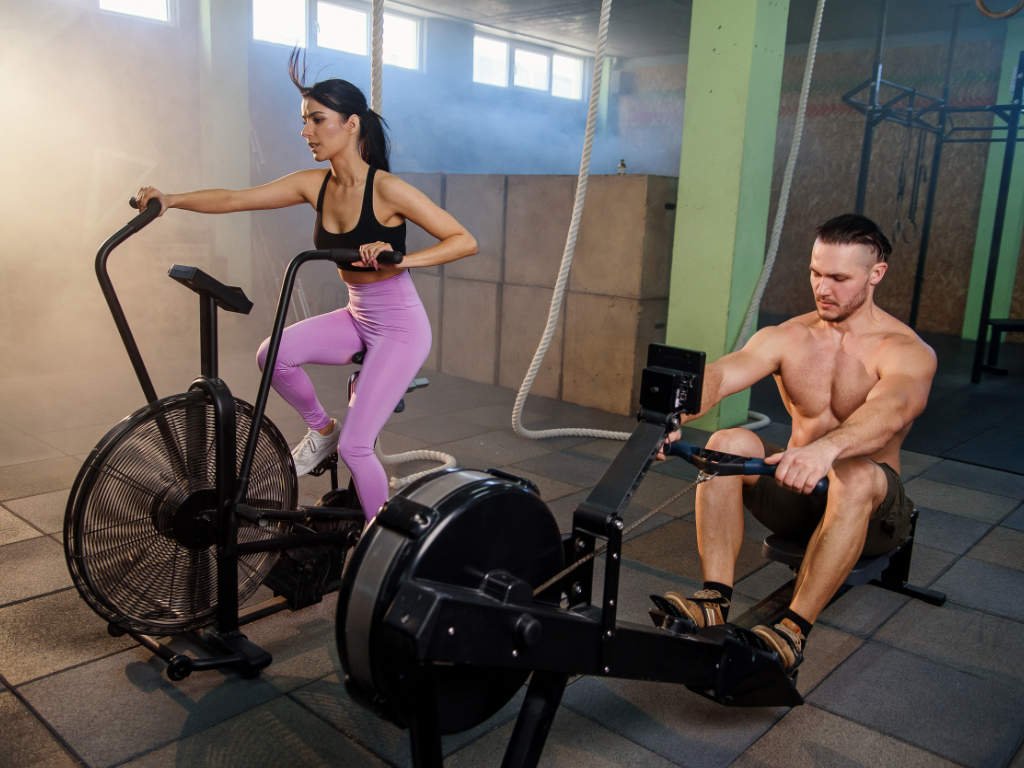 Athletes performing intense cardio exercises on air bike and rowing machine combining cardio with strength training in gym
