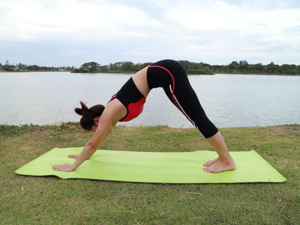 Woman practicing Downward Facing Dog yoga pose for mental clarity and stress relief on green mat by peaceful lake