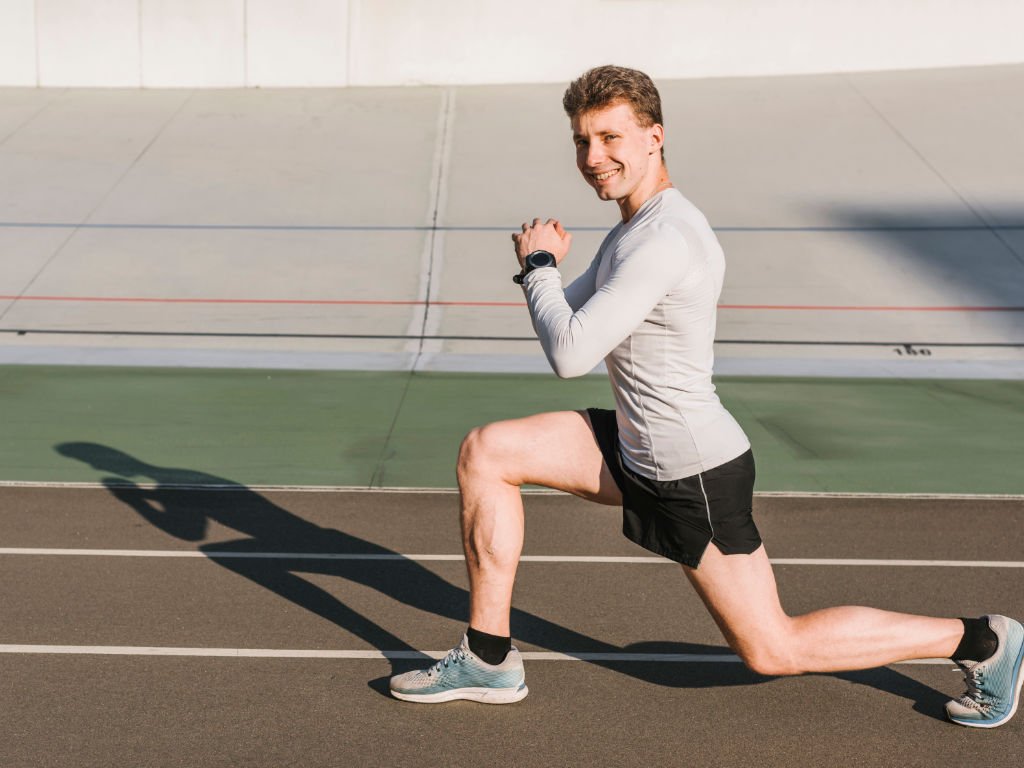 Athletic man performing dynamic stretching exercises with forward lunge movement on outdoor track to enhance workout performance