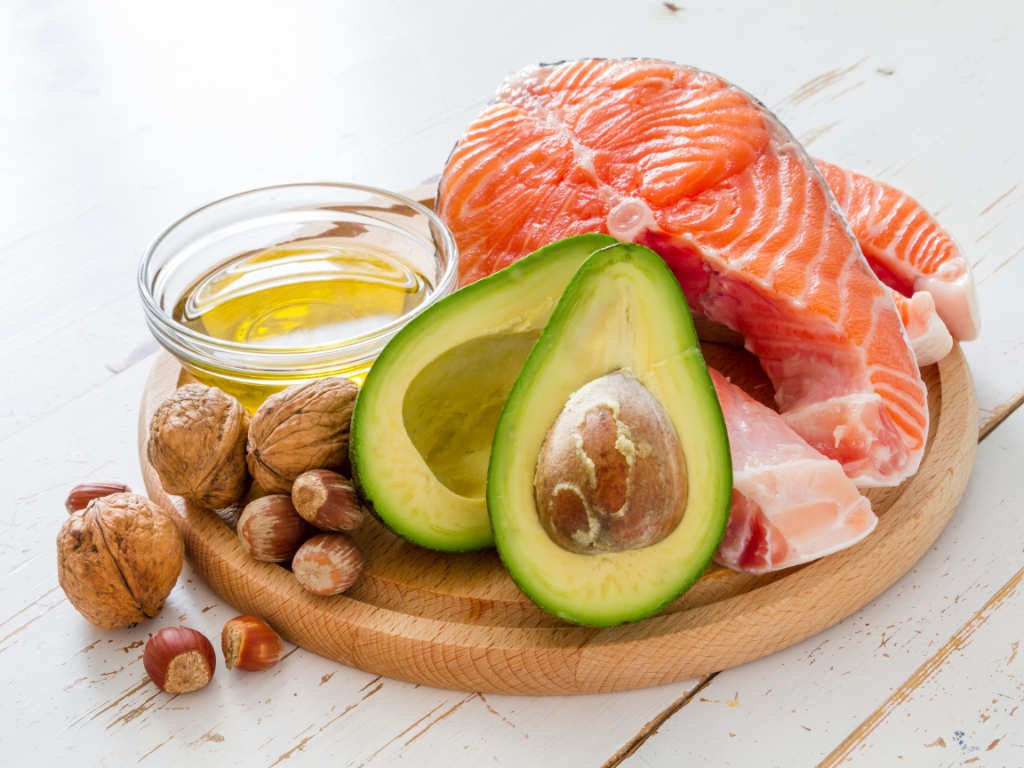 Healthy fats for balanced nutrition including fresh salmon, avocado, mixed nuts, and olive oil arranged on wooden board