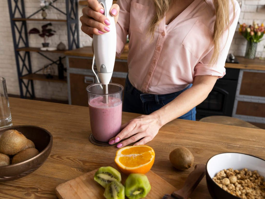 Woman blending smoothies for skin health with fresh kiwi, orange, nuts and other nutrient-dense ingredients on kitchen counter