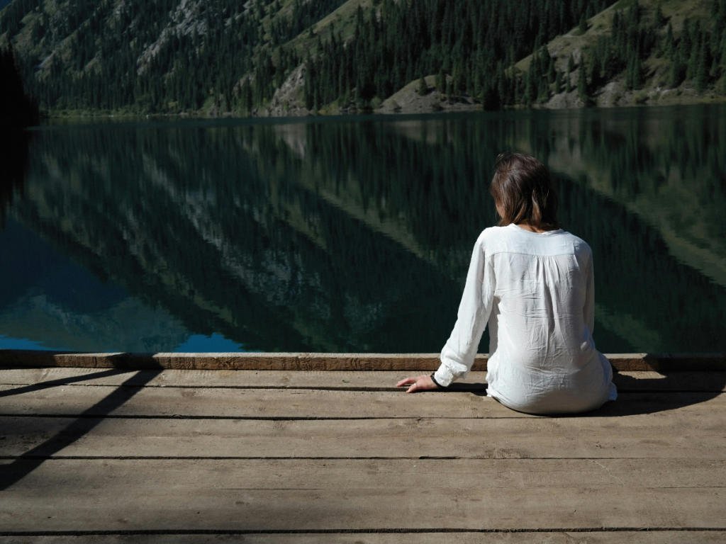Person practicing mindful meditation techniques by serene mountain lake demonstrating science-backed mental health benefits of peaceful reflection