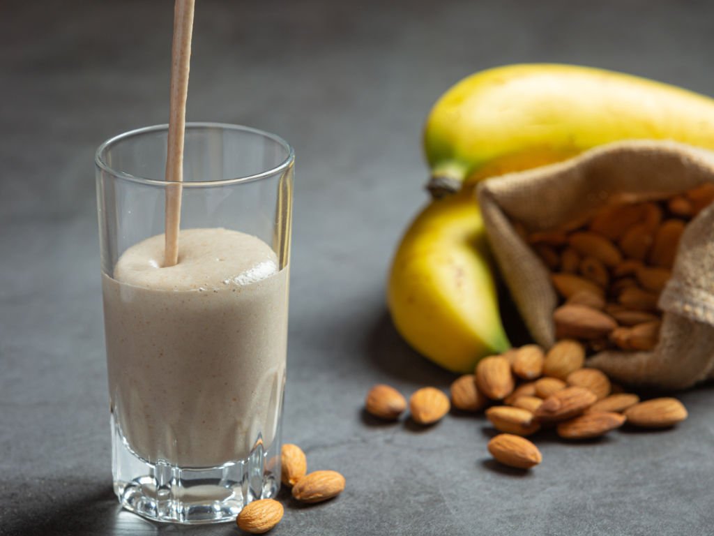Thick creamy meal replacement shake in glass with wooden straw alongside almonds and banana showing balanced nutrition