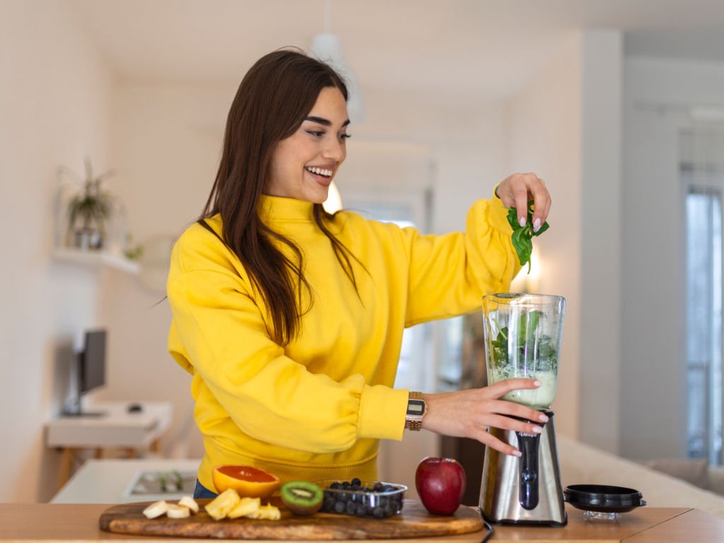 Woman preparing green smoothies for skin health with fresh spinach, kiwi, apple and blueberries in modern kitchen