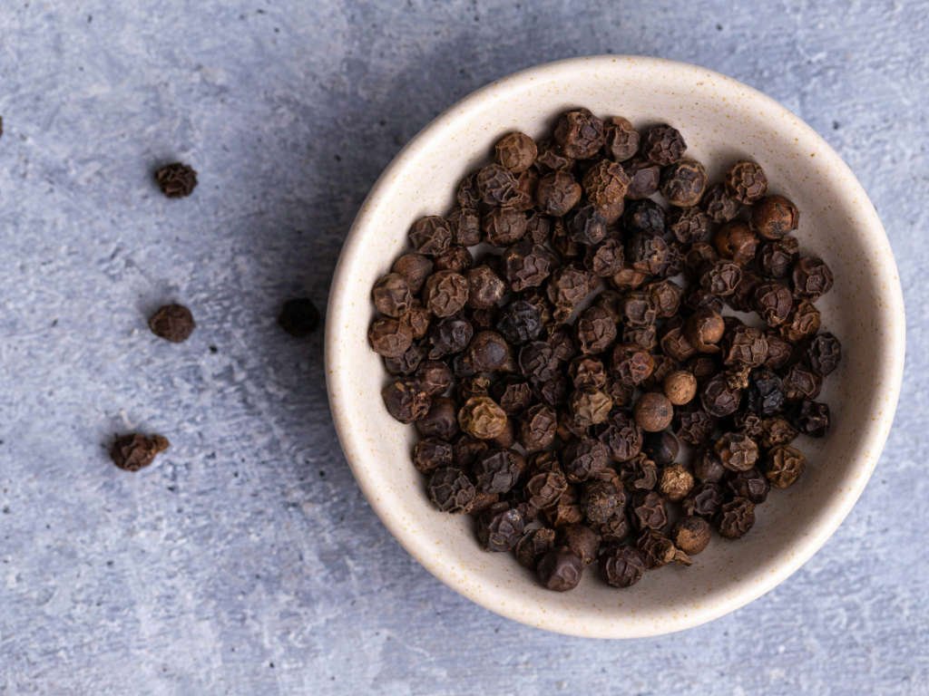 Whole black peppercorns in white bowl on gray surface representing metabolism boosting spices that enhance nutrient absorption and thermogenesis