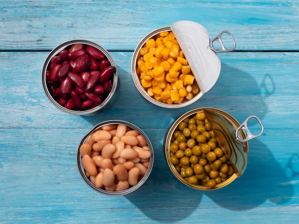 Variety of canned bean staple foods including kidney beans, corn, white beans, and peas in opened cans on blue wooden surface for plant-powered protein meals