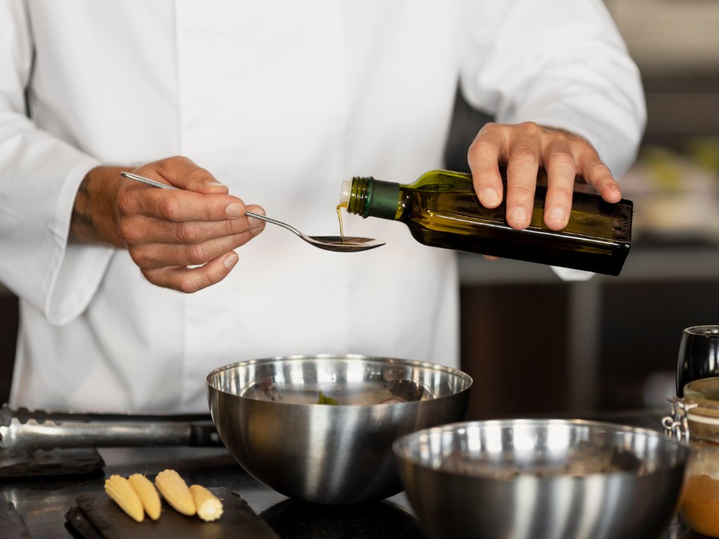 Chef using quality olive oil staple foods while cooking in professional kitchen, drizzling oil from bottle onto spoon for healthy meal preparation