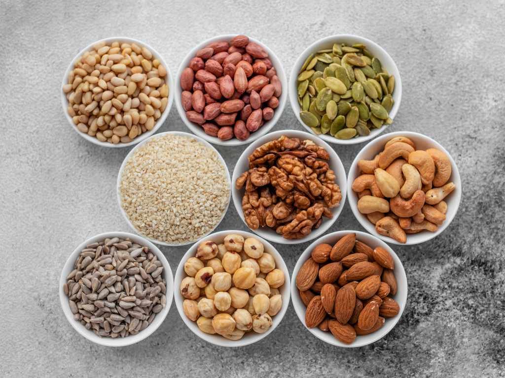 Variety of nuts and seeds staple foods including almonds, walnuts, cashews, sunflower seeds, pumpkin seeds, and sesame seeds arranged in white bowls for healthy meal preparation