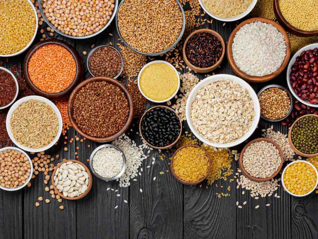 Variety of whole grain staple foods including oats, quinoa, brown rice, lentils, chickpeas, and beans arranged in bowls on wooden surface for healthy meal preparation
