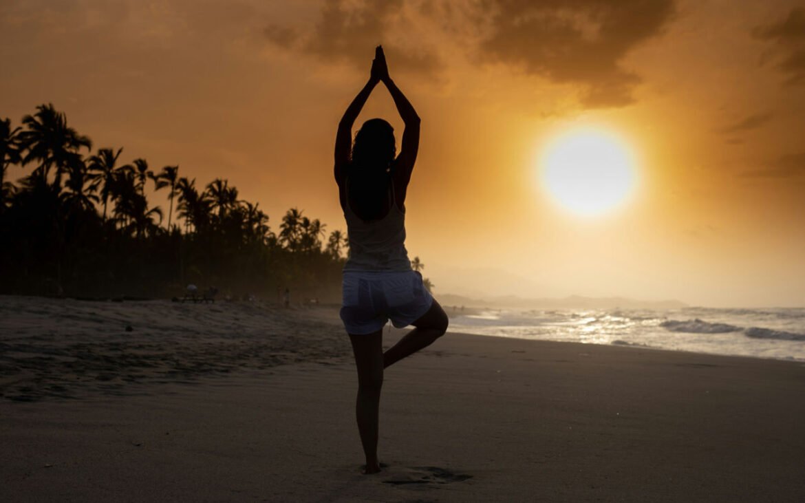 Woman practicing yoga for mental health in tree pose on peaceful beach at sunset with palm trees